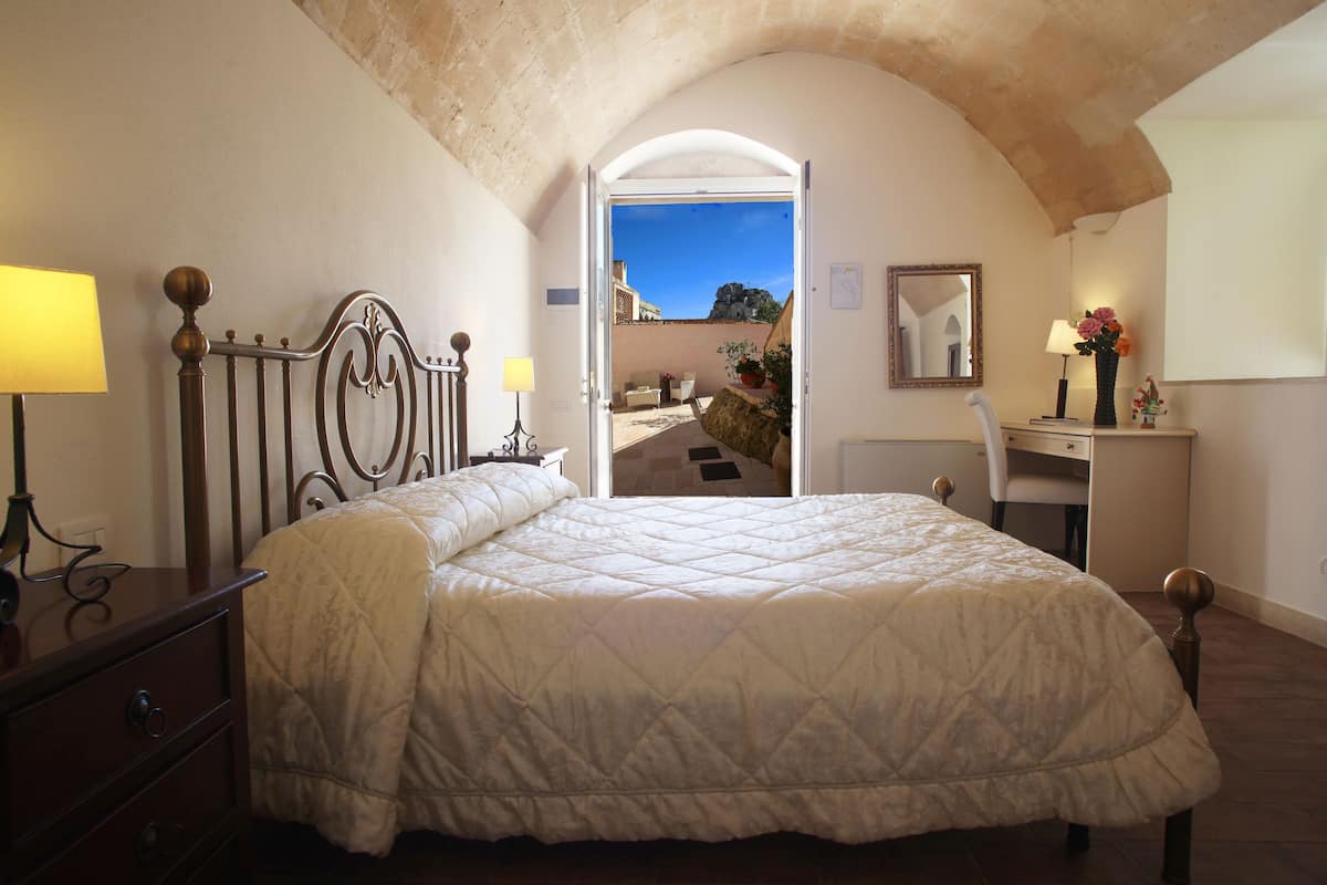 A bedroom with a large bed, a desk, and a chair. There is a door leading to an outdoor area.