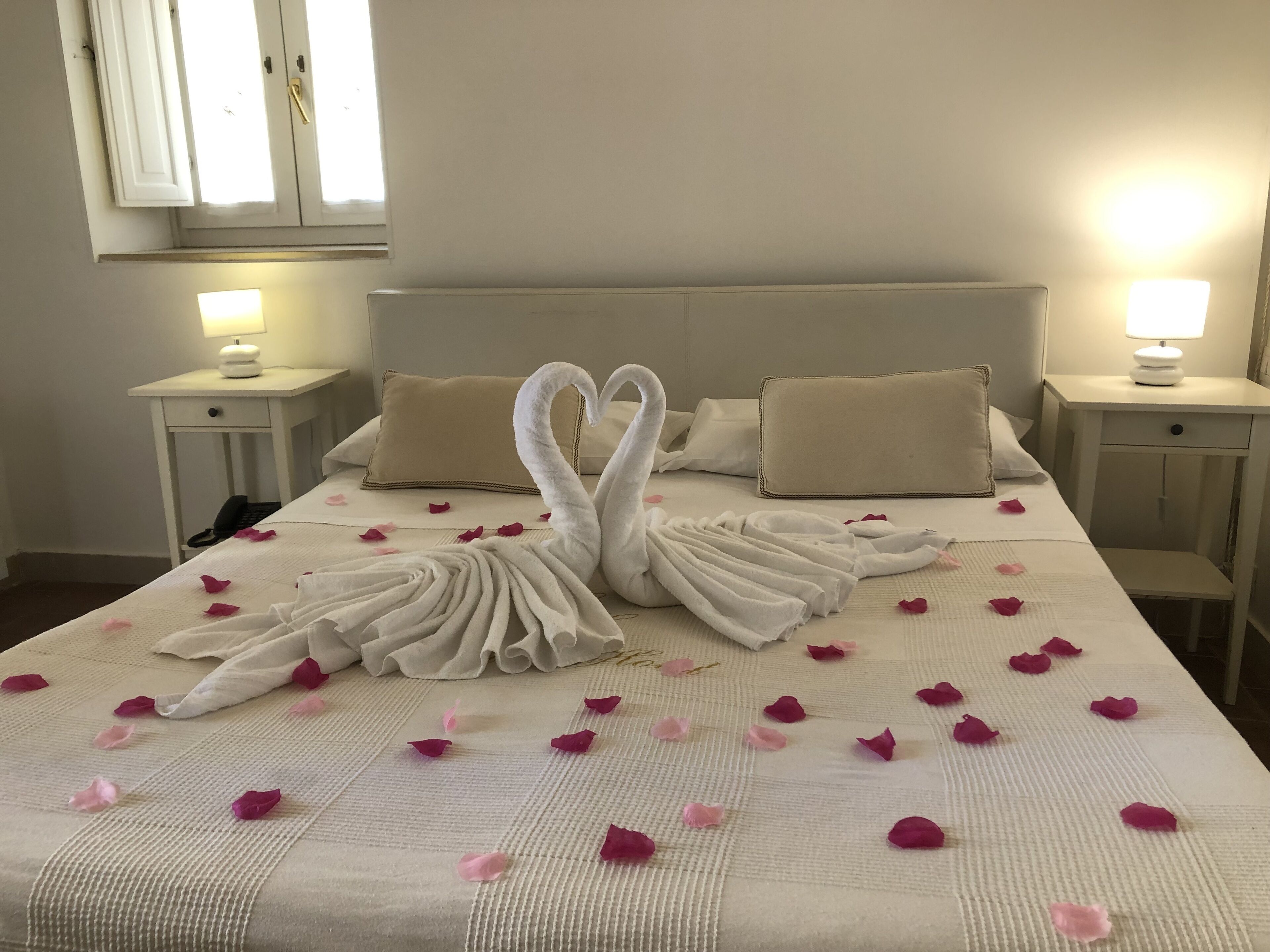 A neatly made bed with a swan towel design and rose petals, two bedside lamps, and a window with curtains.