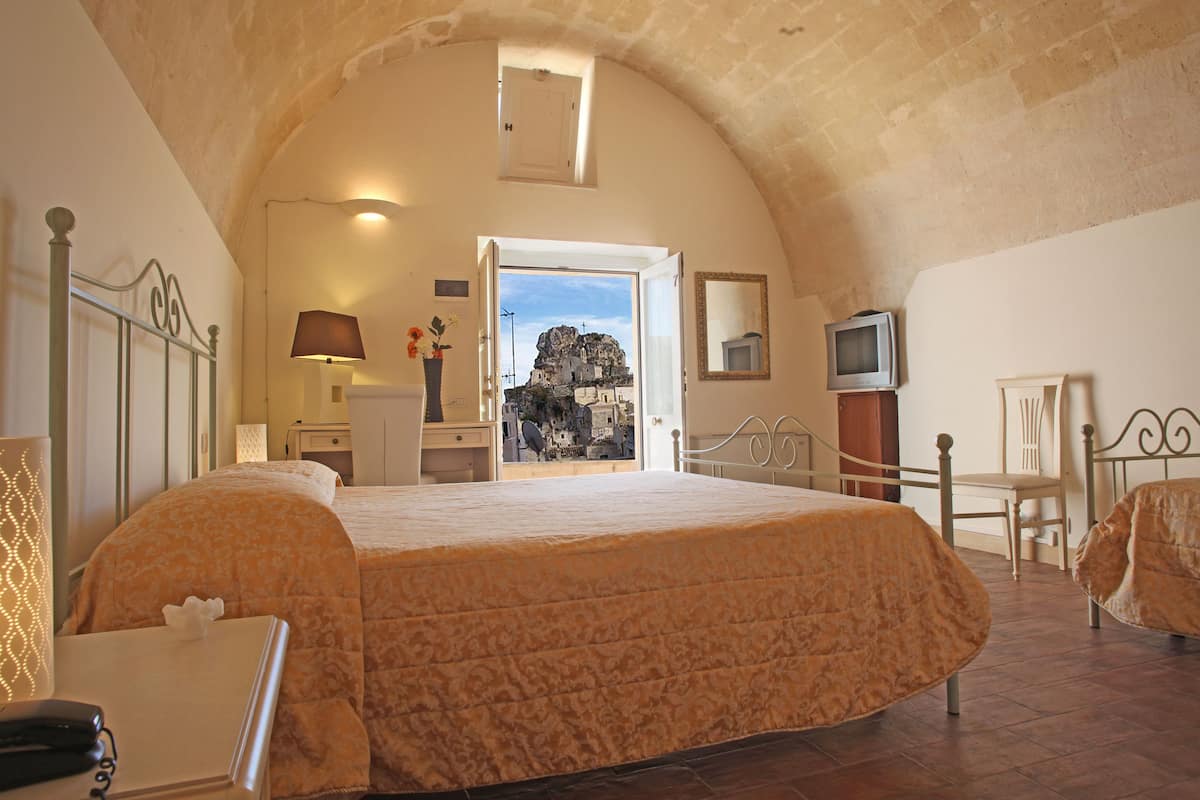 A bedroom with a large bed, a desk, a chair, a TV, and a view of a rocky landscape through an open door.