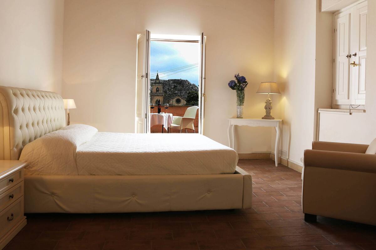 A bedroom with a bed, nightstand, chair, and a view of a building through an open door.