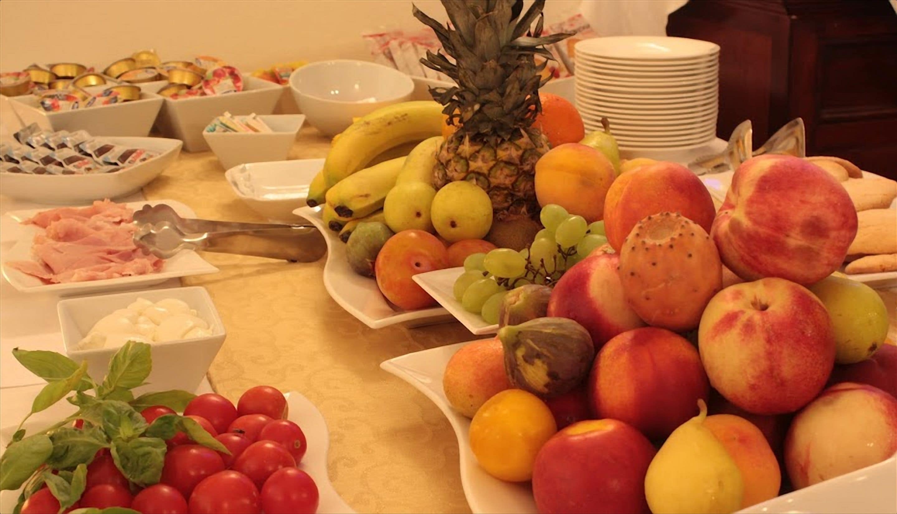 Una mesa de buffet con diversas frutas, una piña, fuentes de comida y una pila de platos.