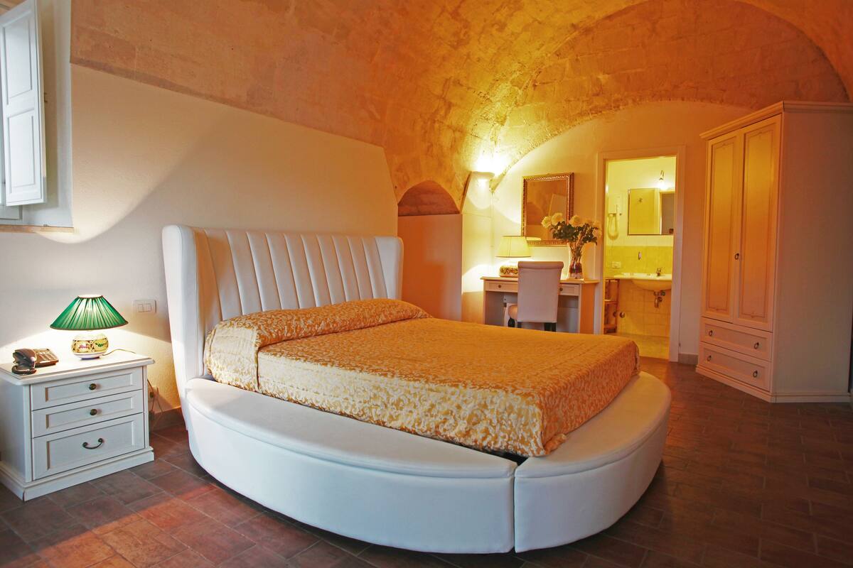A bedroom with a curved bed, a nightstand with drawers, a lamp, and a bathroom visible in the background.