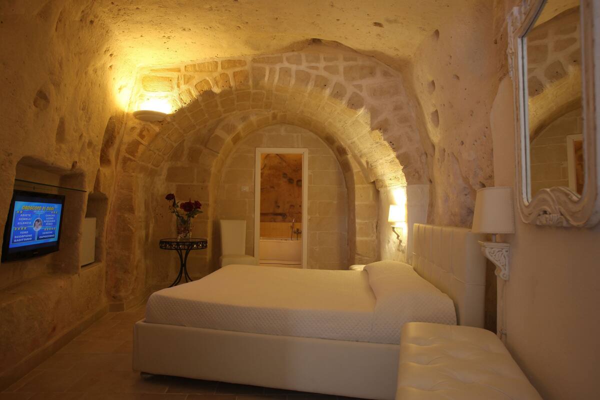 A bedroom with a stone archway, a bed, a television, and a bathroom visible through the archway.