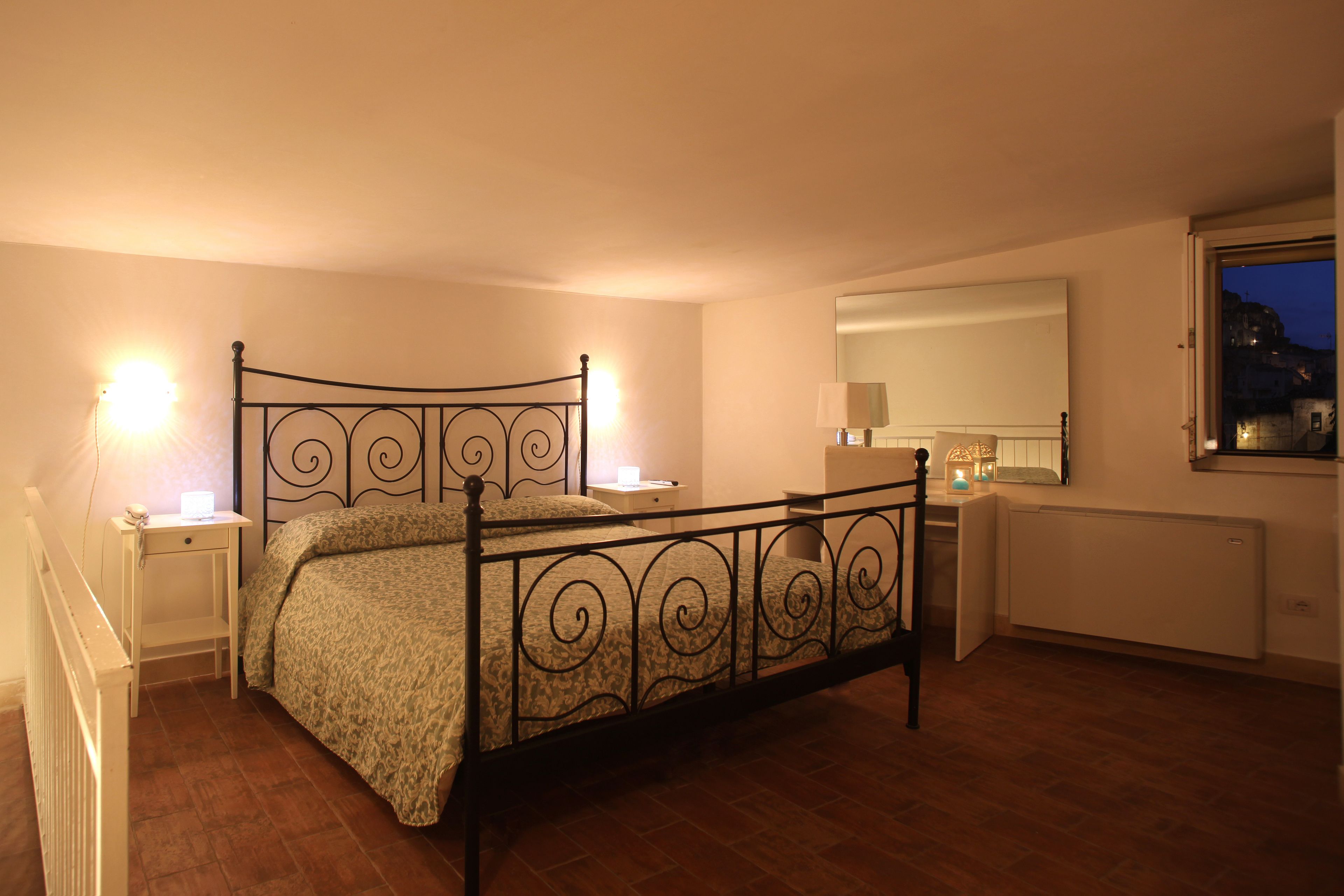 Quarto com estrutura de cama em metal, mesa de cabeceira com abajur, um criado-mudo, um espelho e janela com vista para o exterior.