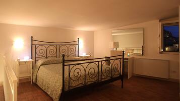 Quarto com estrutura de cama em metal, mesa de cabeceira com abajur, um criado-mudo, um espelho e janela com vista para o exterior.