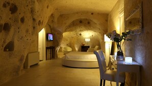 A room with a round bed, a chair, a small table with a vase, and a TV on a stand. - Caveoso Hotel (Matera)
