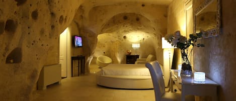 A room with a round bed, a chair, a small table with a vase, and a TV on a stand.