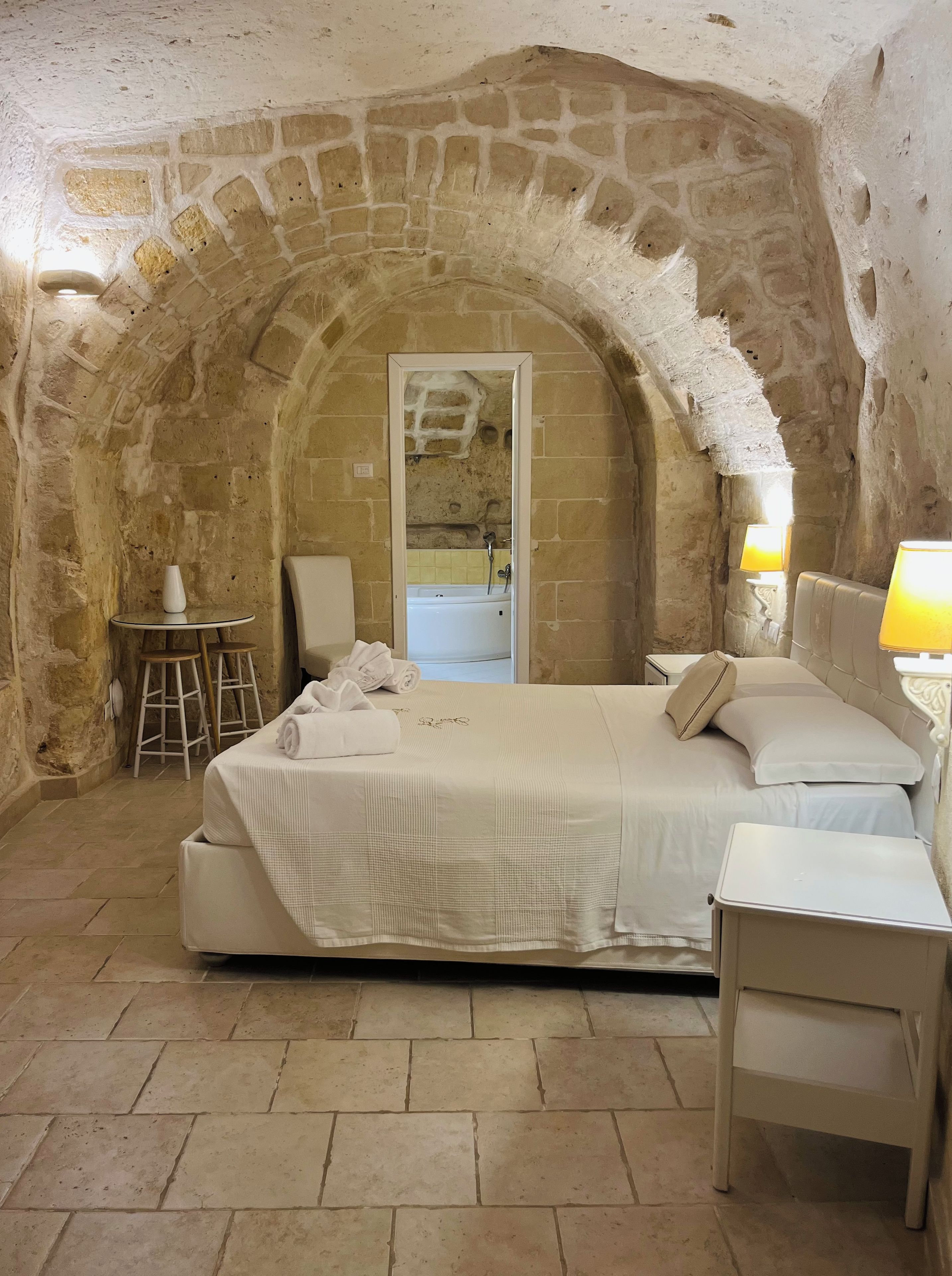 A room with a stone archway, a bed with white linens, a small round table with a white vase, and a nightstand with a lamp.