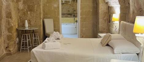 A room with a stone archway, a bed with white linens, a small round table with a white vase, and a nightstand with a lamp.