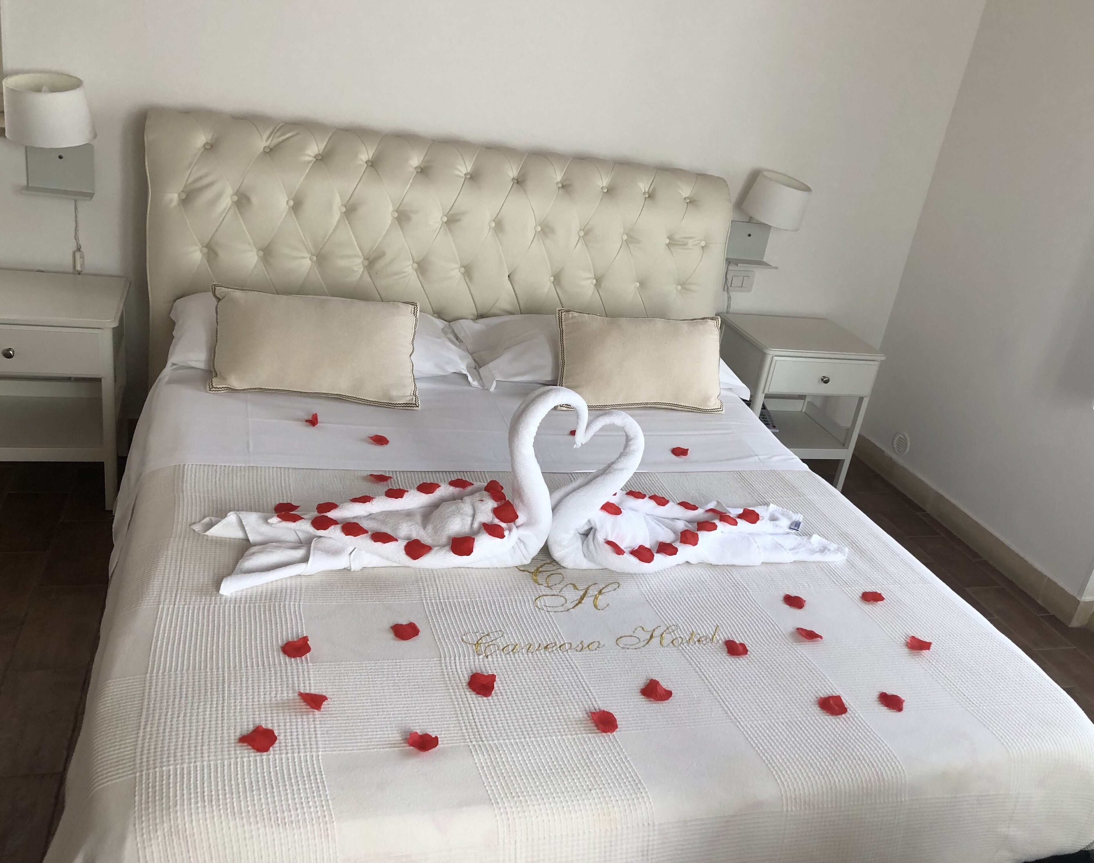 A neatly made bed with a swan-shaped towel arrangement and rose petals, a tufted headboard, and bedside lamps.