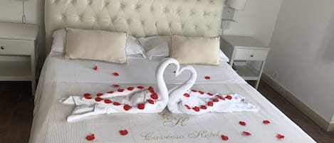 Een net opgemaakt bed met een zwaan van handdoeken en rozenblaadjes, een gestoffeerd hoofdeinde en bedlampjes.