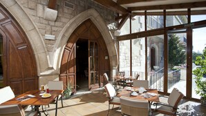 Un amplio comedor al aire libre con paredes de piedra y vigas de madera, con mesas y sillas, y vistas a la calle a través de grandes ventanales.
