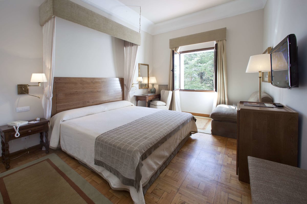 Habitación de hotel con una cama grande, mesitas de noche con lámparas, un escritorio, una silla, un televisor y una ventana con cortinas.