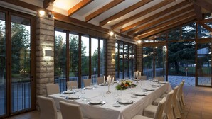 Una larga mesa de comedor preparada para una comida, con sillas, un pilar de piedra y ventanas grandes que dan a un paisaje de árboles.