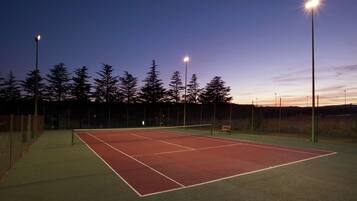 Un court de tennis en plein air, éclairé par des projecteurs, au crépuscule, entouré d’arbres et agrémenté d’un banc.