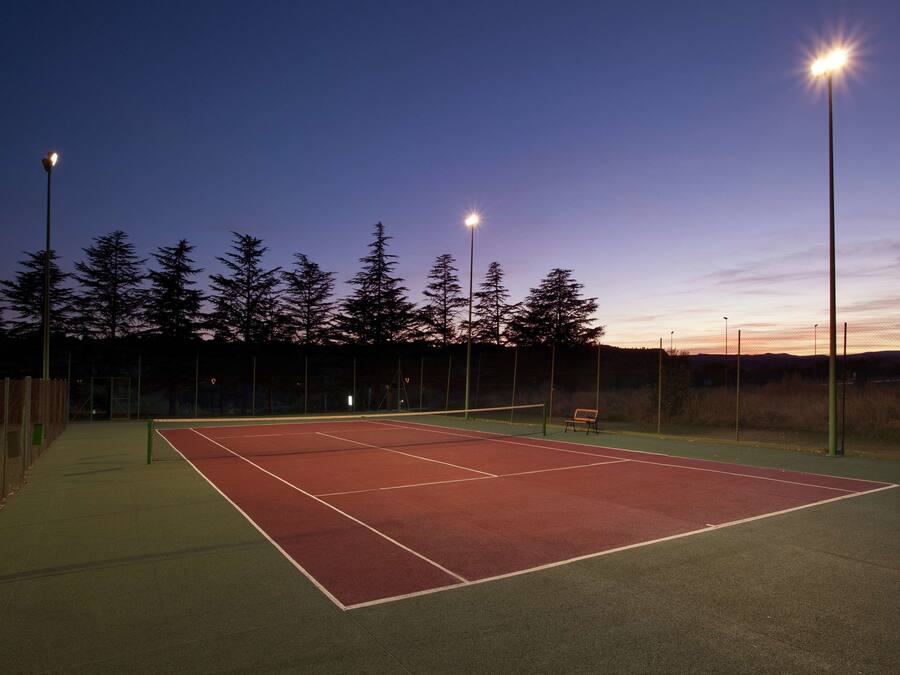 Un court de tennis en plein air, éclairé par des projecteurs, au crépuscule, entouré d’arbres et agrémenté d’un banc.