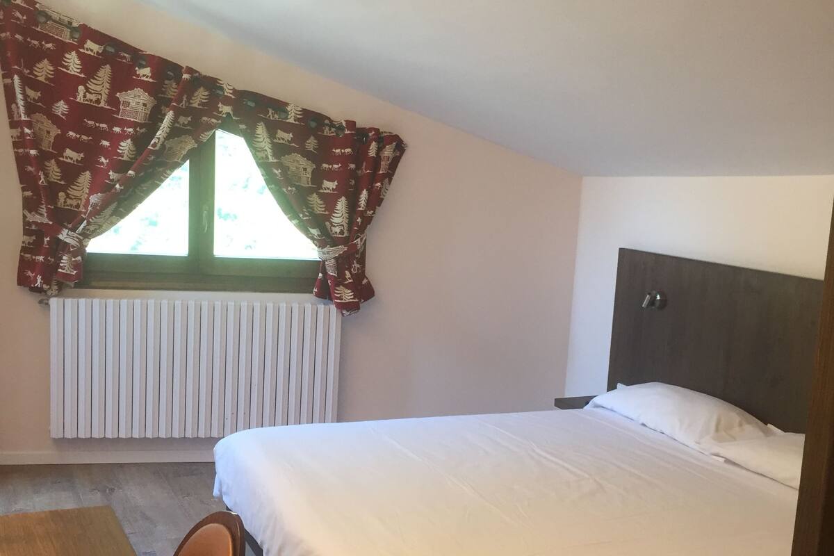 A bedroom with a bed, a window with curtains, a radiator, a wooden table, and a chair.