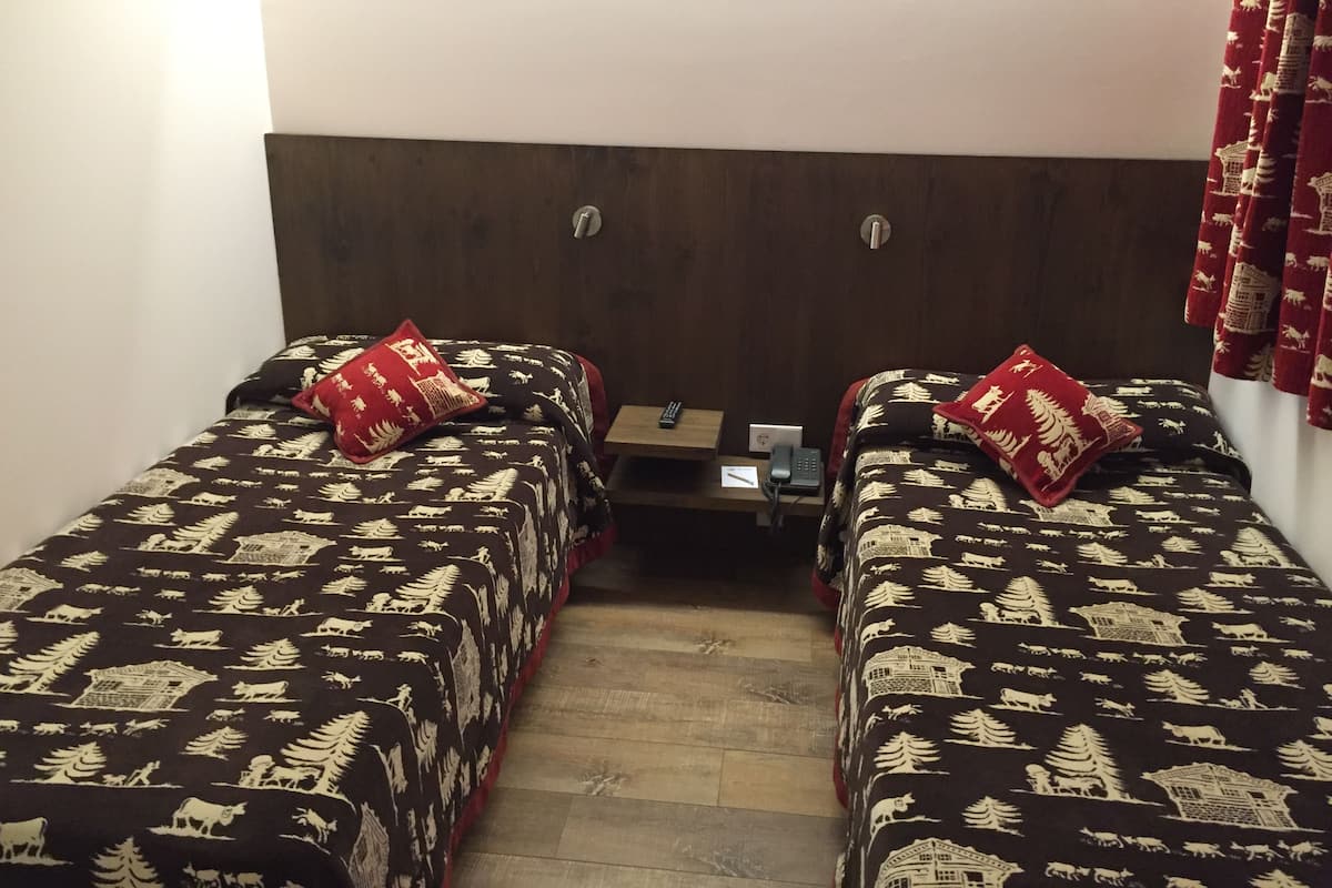 A room with two beds, each with a patterned bedspread and a red pillow. There is a small table with a phone and a power outlet between the beds.