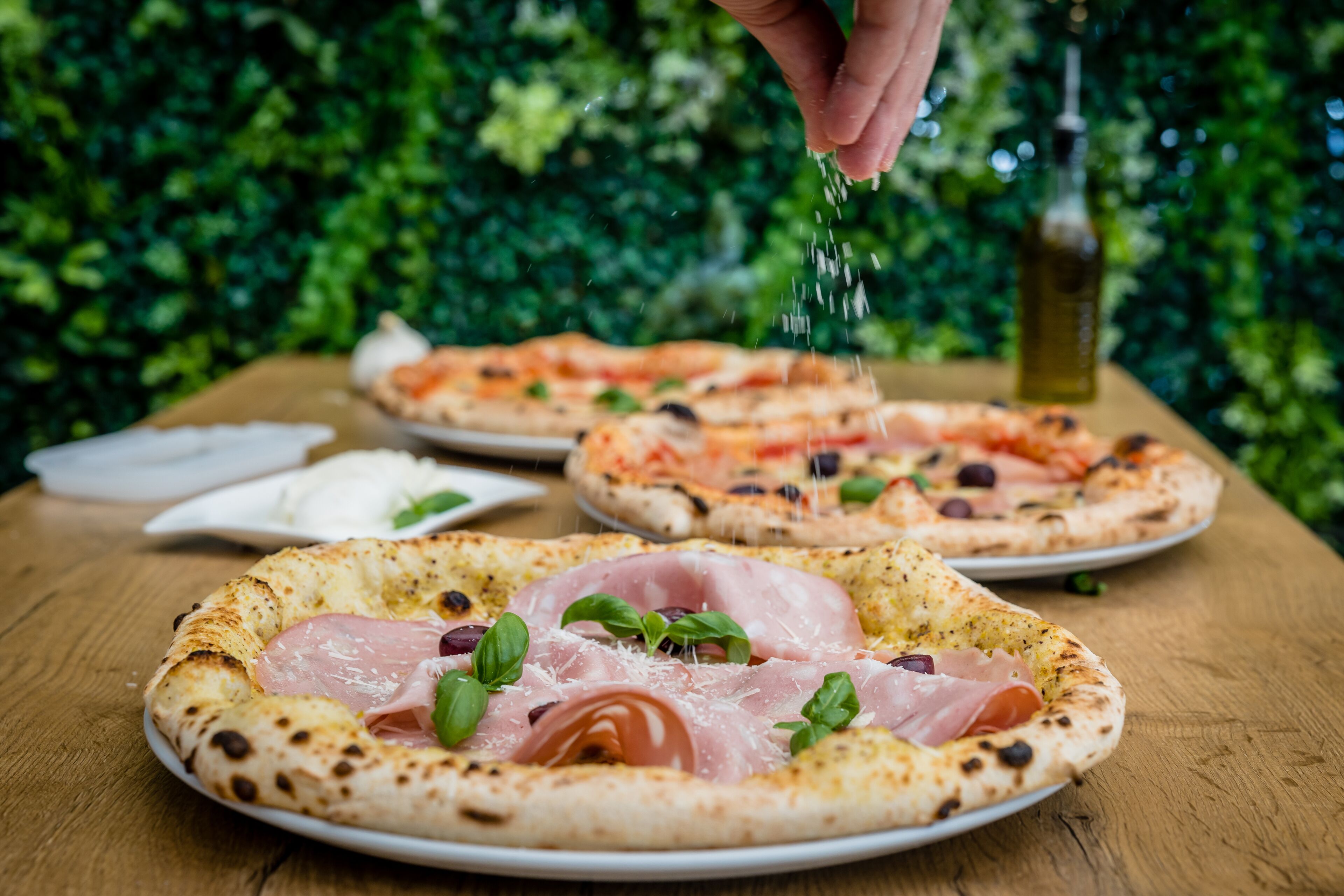 Ein Holztisch mit drei Pizzen, eine belegt mit Basilikum und Schinken, eine weitere mit Oliven und Tomaten sowie eine dritte mit verschiedenen Belägen.