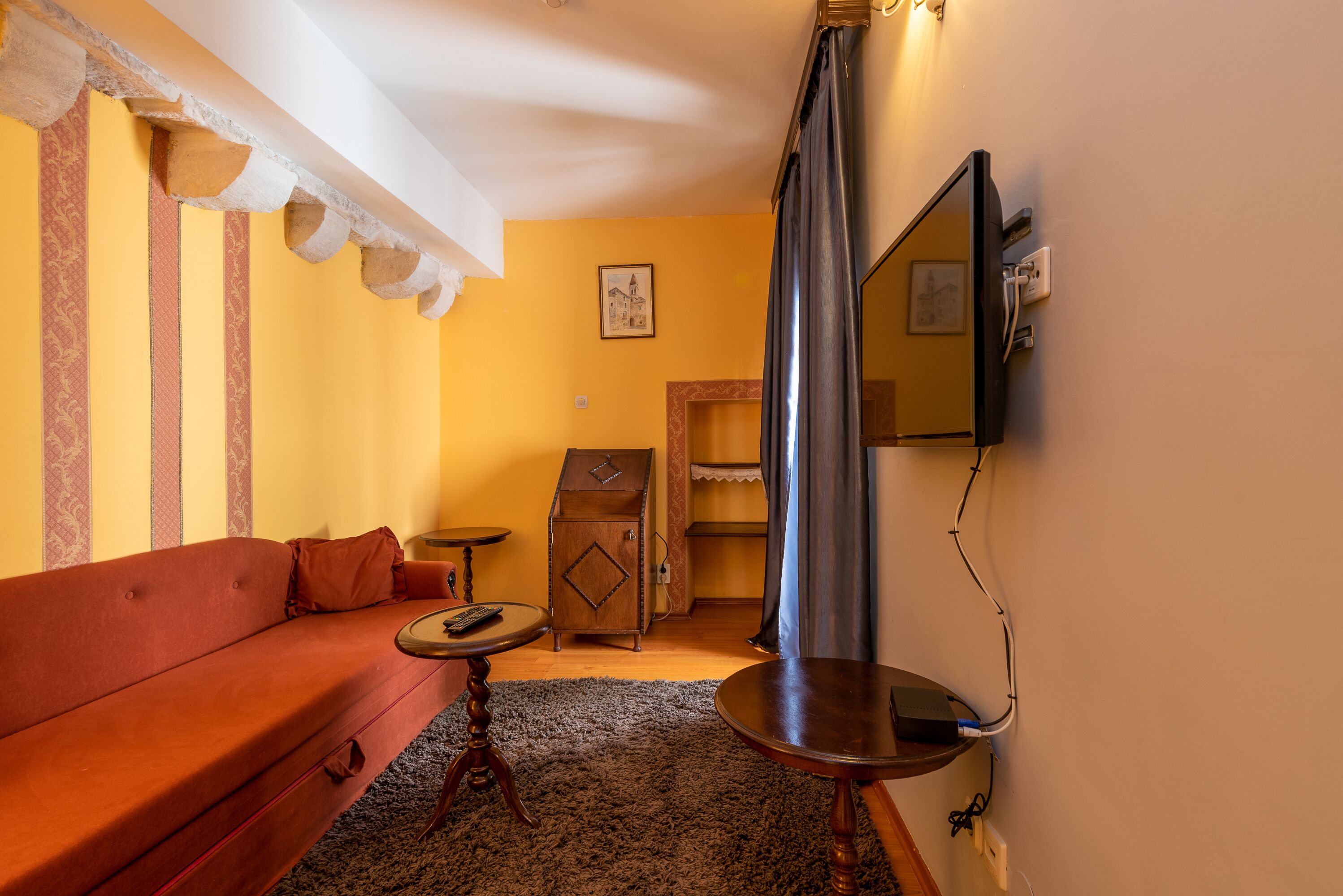 Una habitación acogedora con un sofá rojo, un mueble de madera pequeño, una mesa redonda y una cortina colgada.