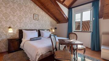 Un dormitorio con techo de madera, una cama con ropa de cama blanca, una mesita con una silla y un ventanal con cortinas.