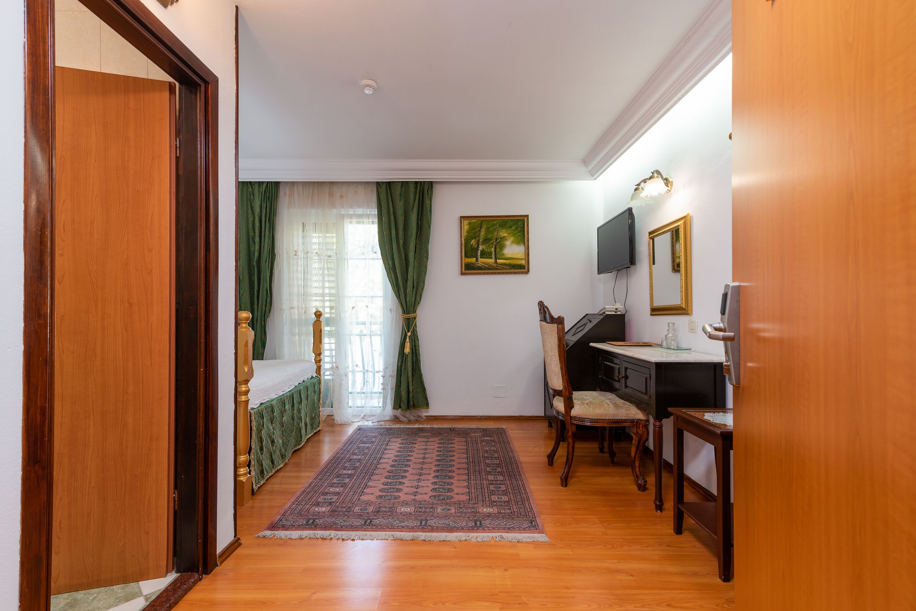 A room with a wooden floor, a bed with green curtains, a desk with a chair, and a painting on the wall.
