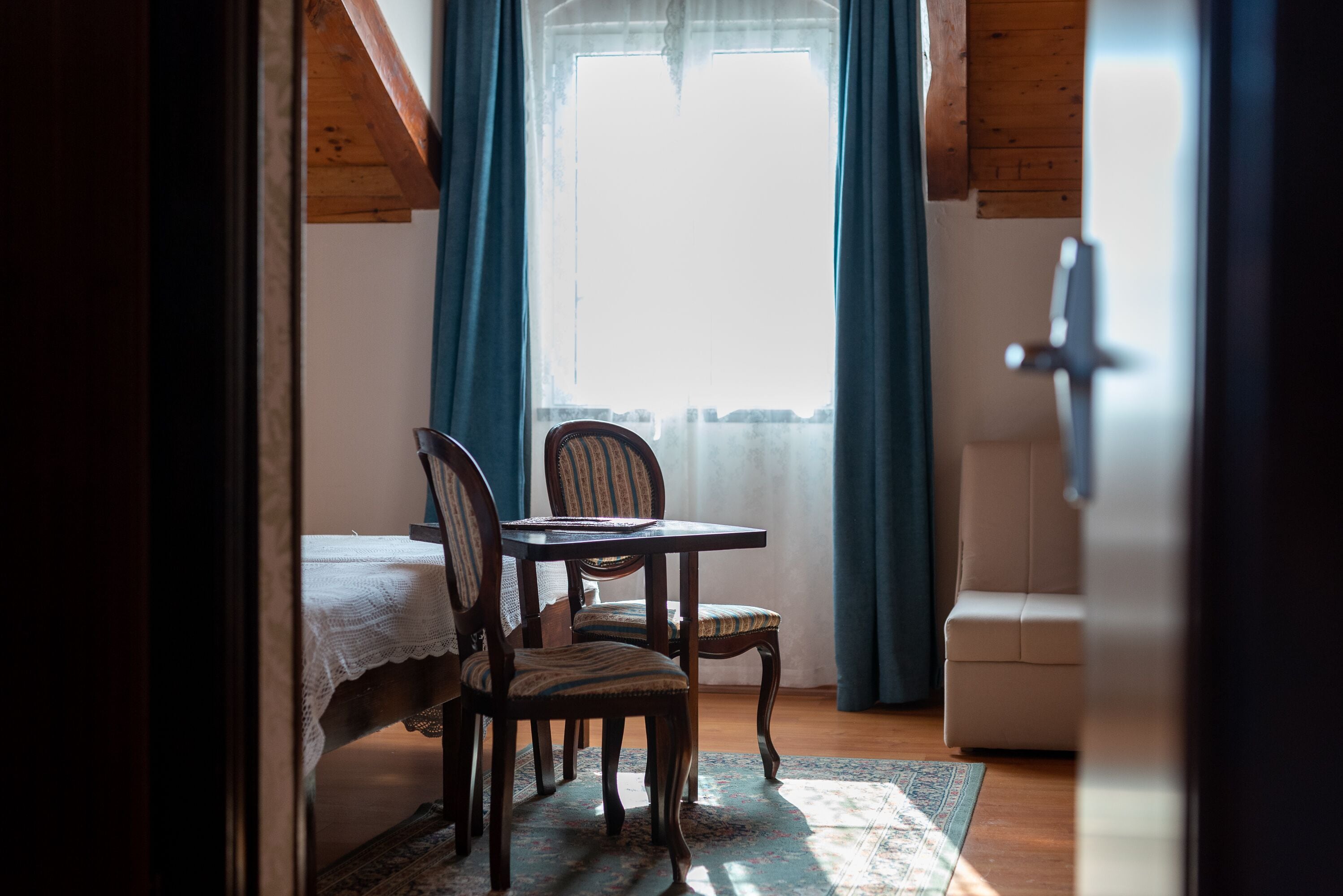A cozy room with a wooden ceiling, a dining table with chairs, a bed with a white blanket, and a window with blue curtains.