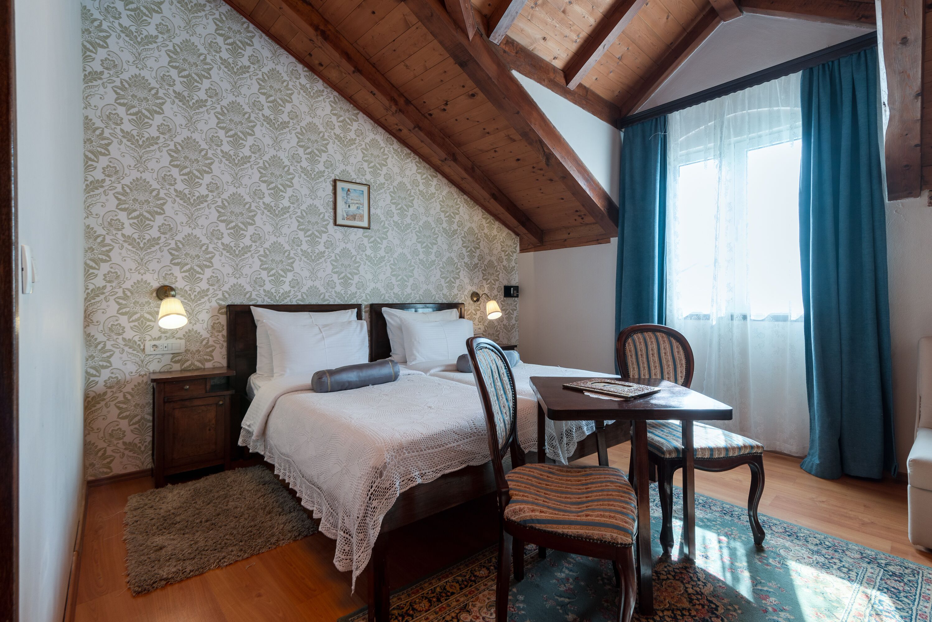 Un dormitorio con techo de madera, una cama con sábanas blancas, una mesita de noche, una mesa con una silla y una ventana con cortinas.
