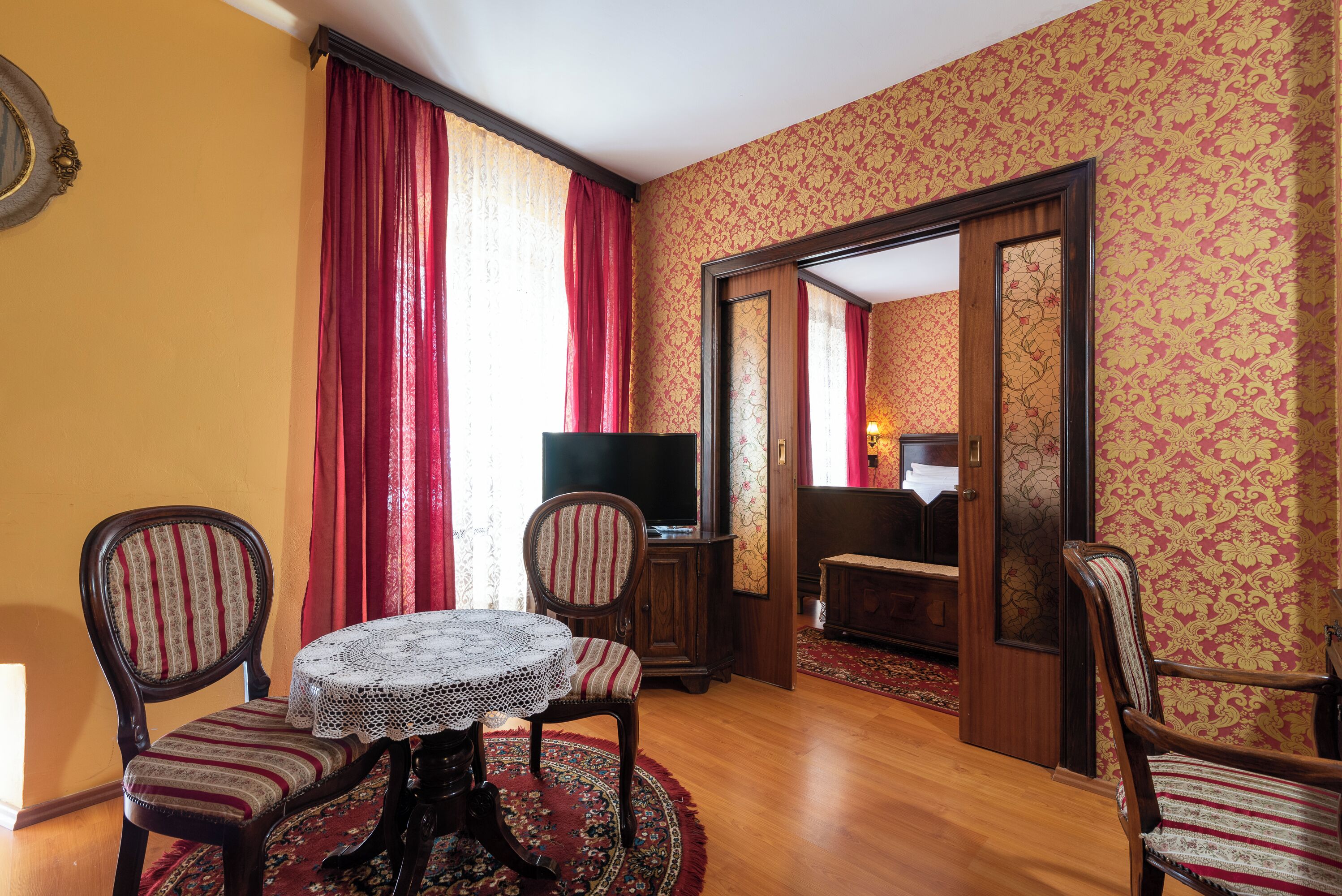 A room with a wooden floor, a round table with a lace tablecloth, two striped chairs, a television, and a mirror reflecting a room with a red curtain and patterned wallpaper.