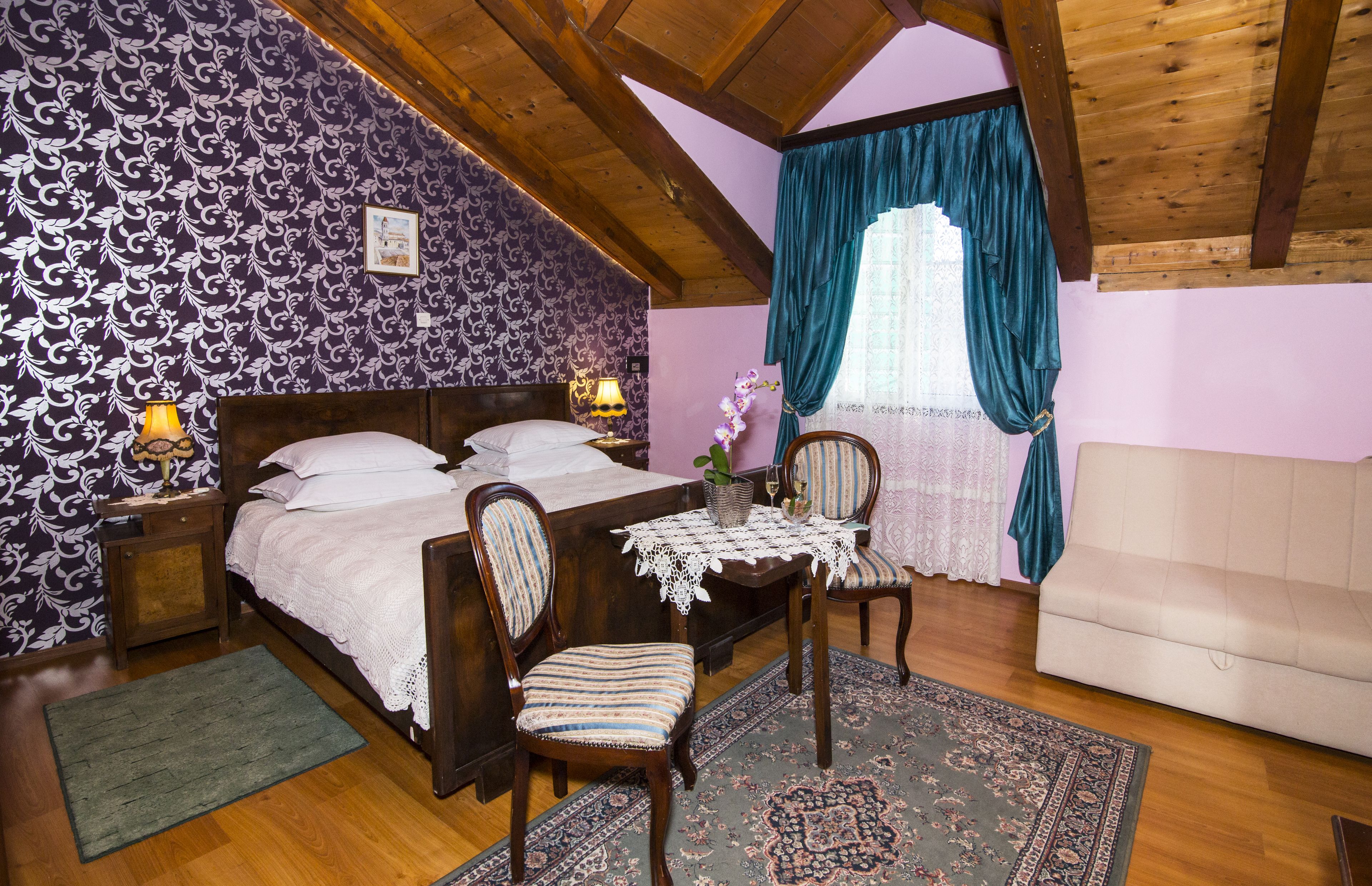 Un dormitorio con techo de madera, una cama con cubrecama blanco, dos sillas, una mesita con mantel de encaje y una ventana con cortinas color turquesa.