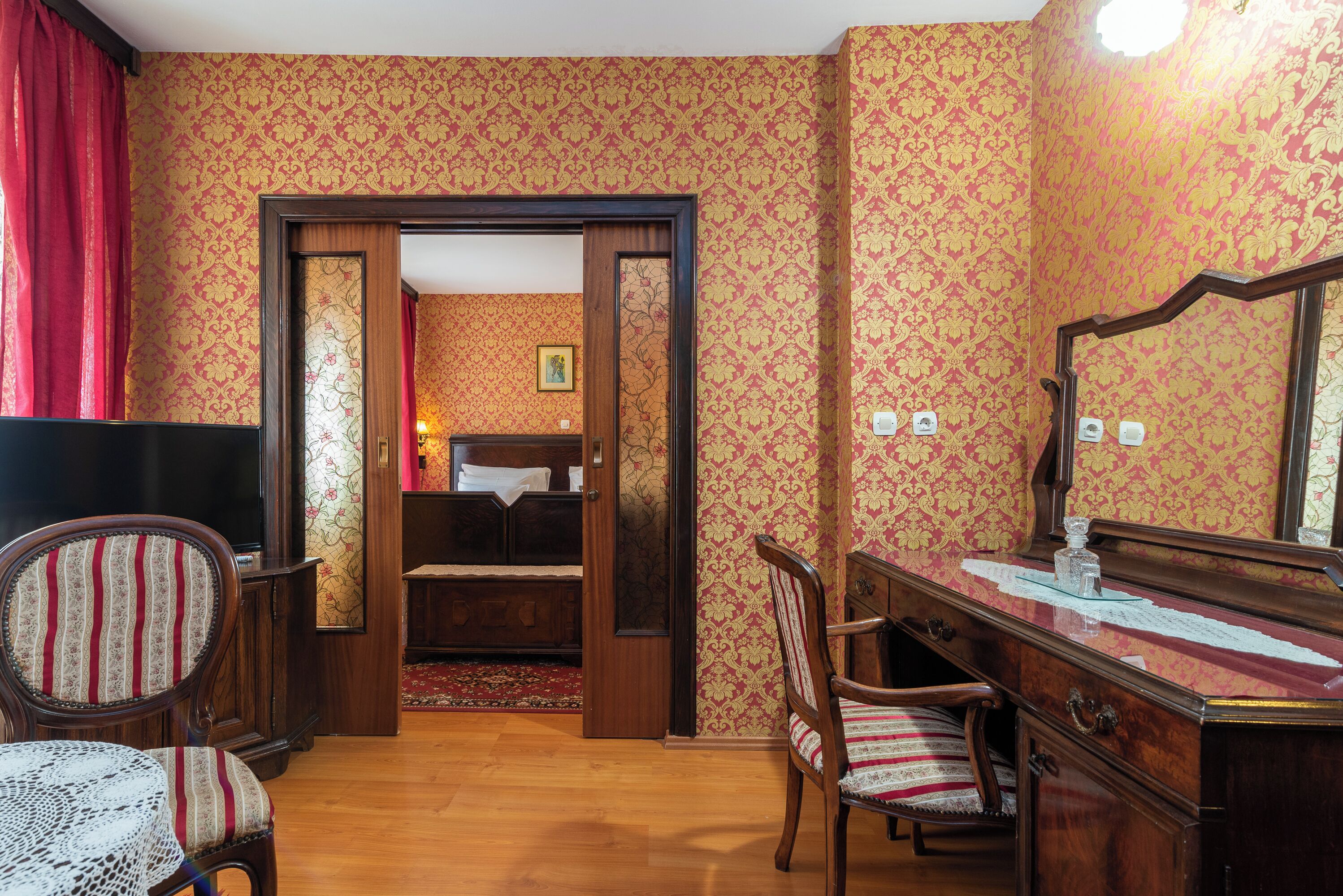 A room with patterned wallpaper, a wooden desk with a mirror, a chair, and a bed with a red curtain.