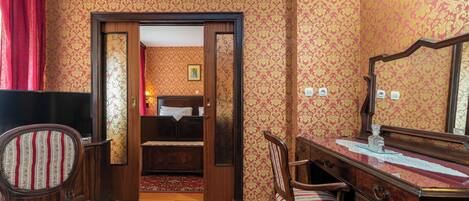 A room with patterned wallpaper, a wooden desk with a mirror, a chair, and a bed with a red curtain.