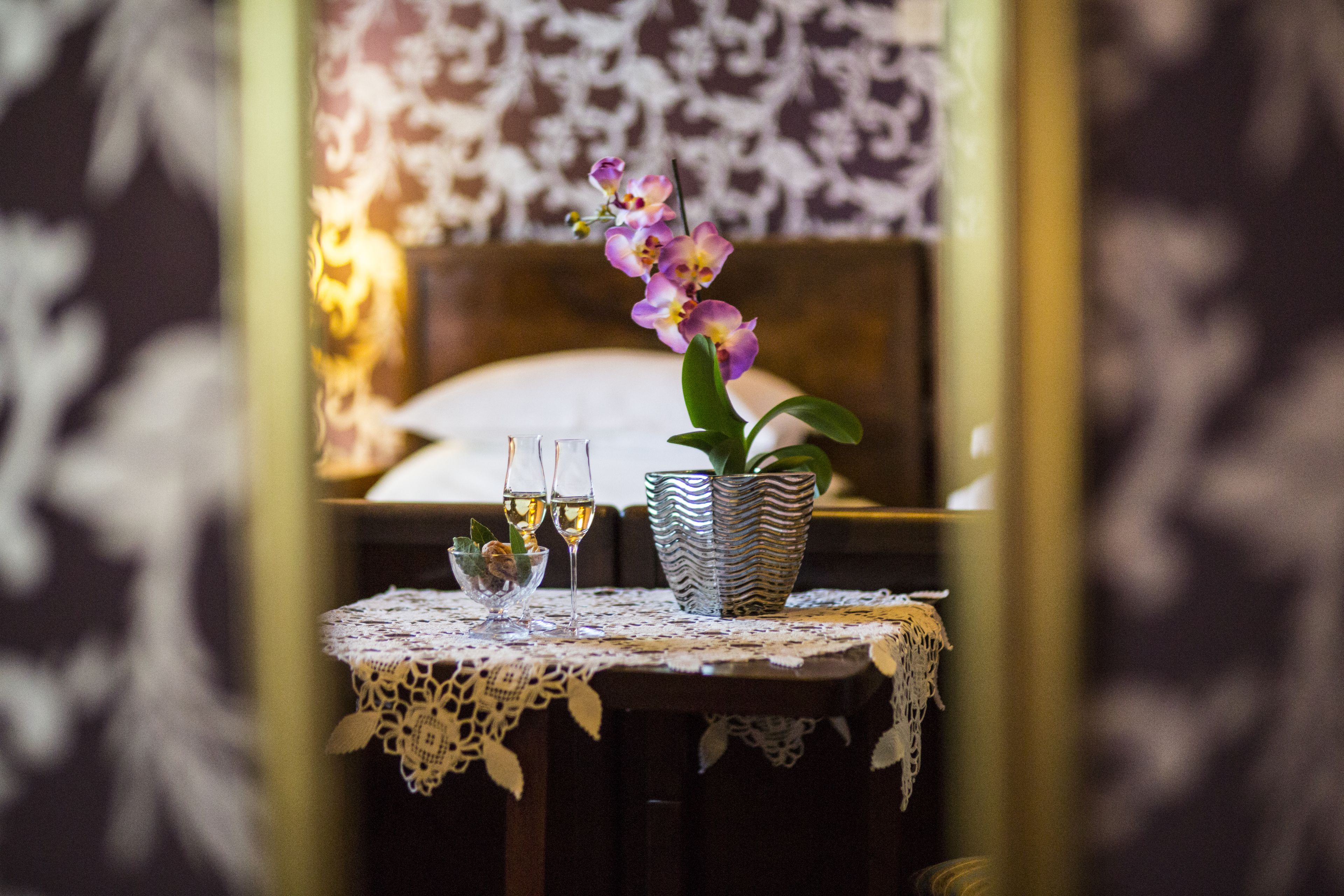 A table with a lace tablecloth, two wine glasses, a small vase with a plant, and a patterned wall in the background.