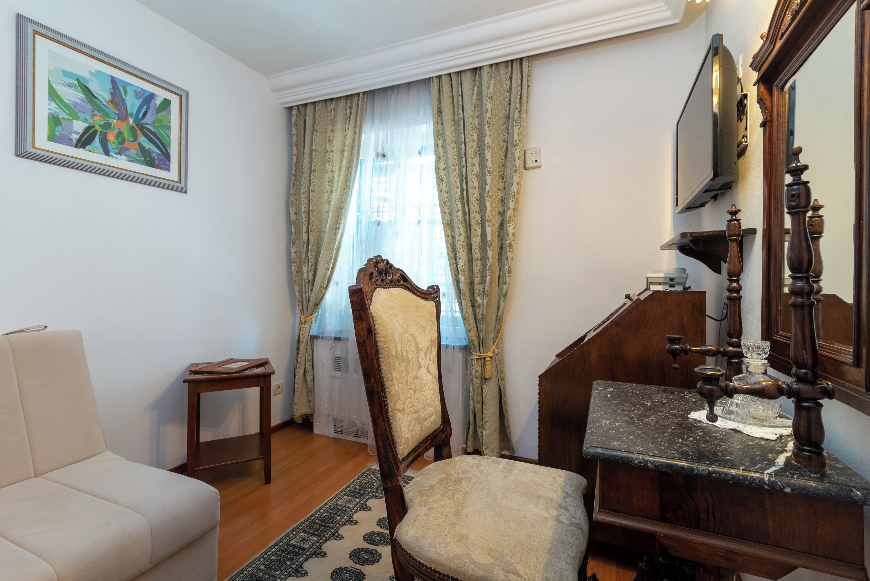 A room with a wooden chair, a small table, a sofa, a mirror, and a painting on the wall.