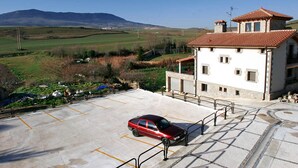 A building with a red-tiled roof, a parking lot with a red car, and a view of green fields and hills in the background. - Agorreta (Galar)