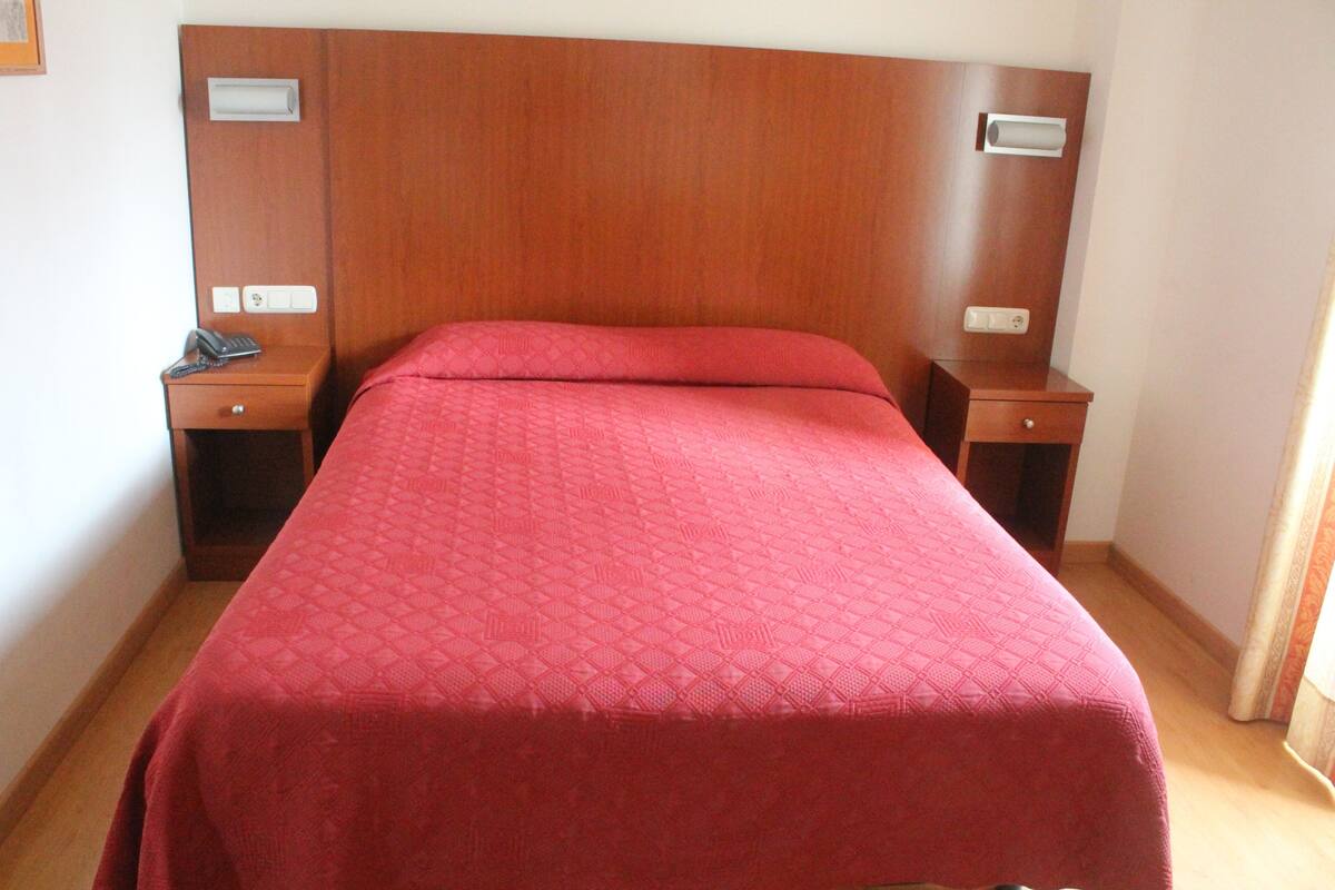 A hotel room with a wooden headboard, a red bedspread, two bedside tables with lamps, and a single bedside drawer.