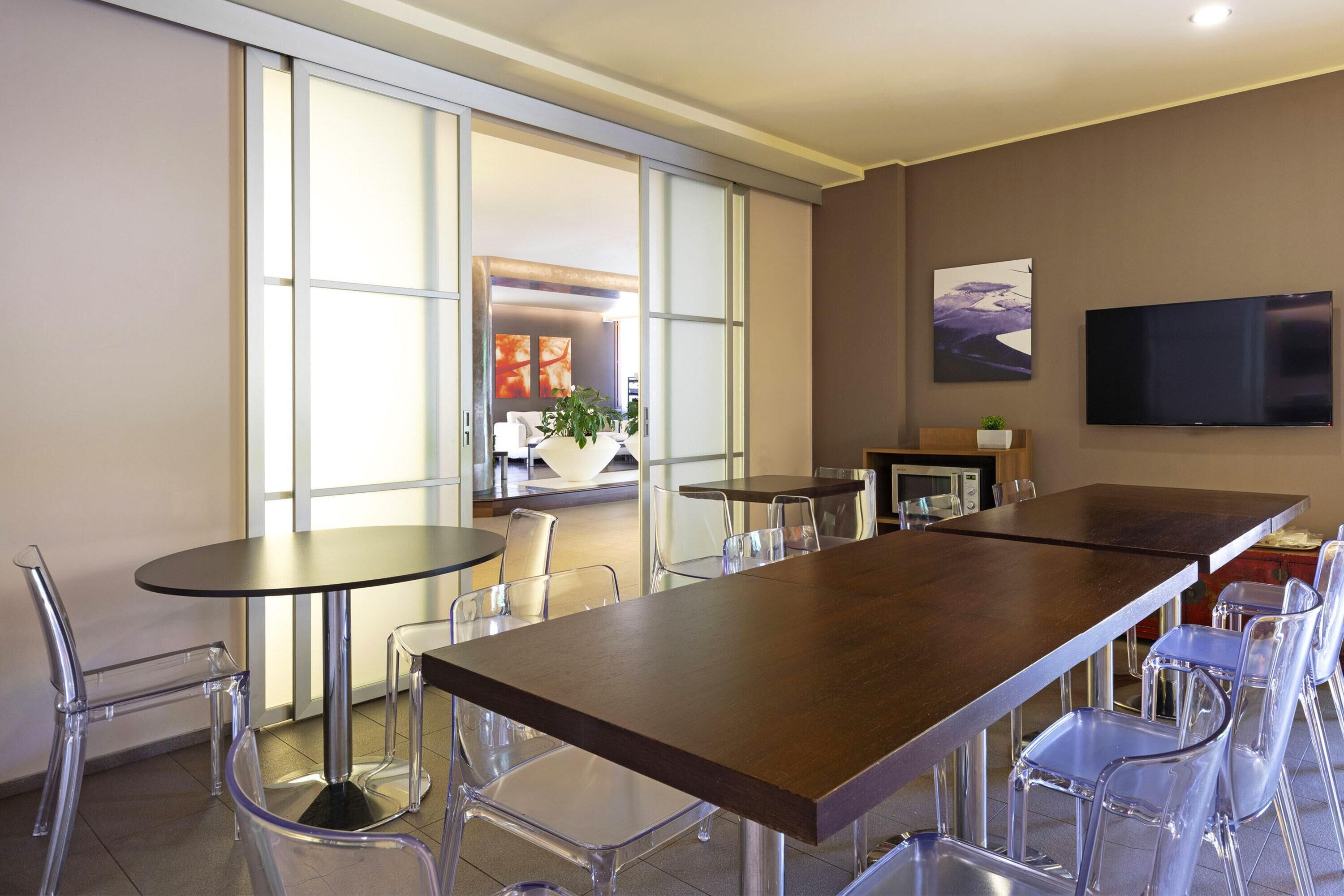 A modern dining area with a long table and clear chairs, large windows, and a flat-screen TV on the wall.