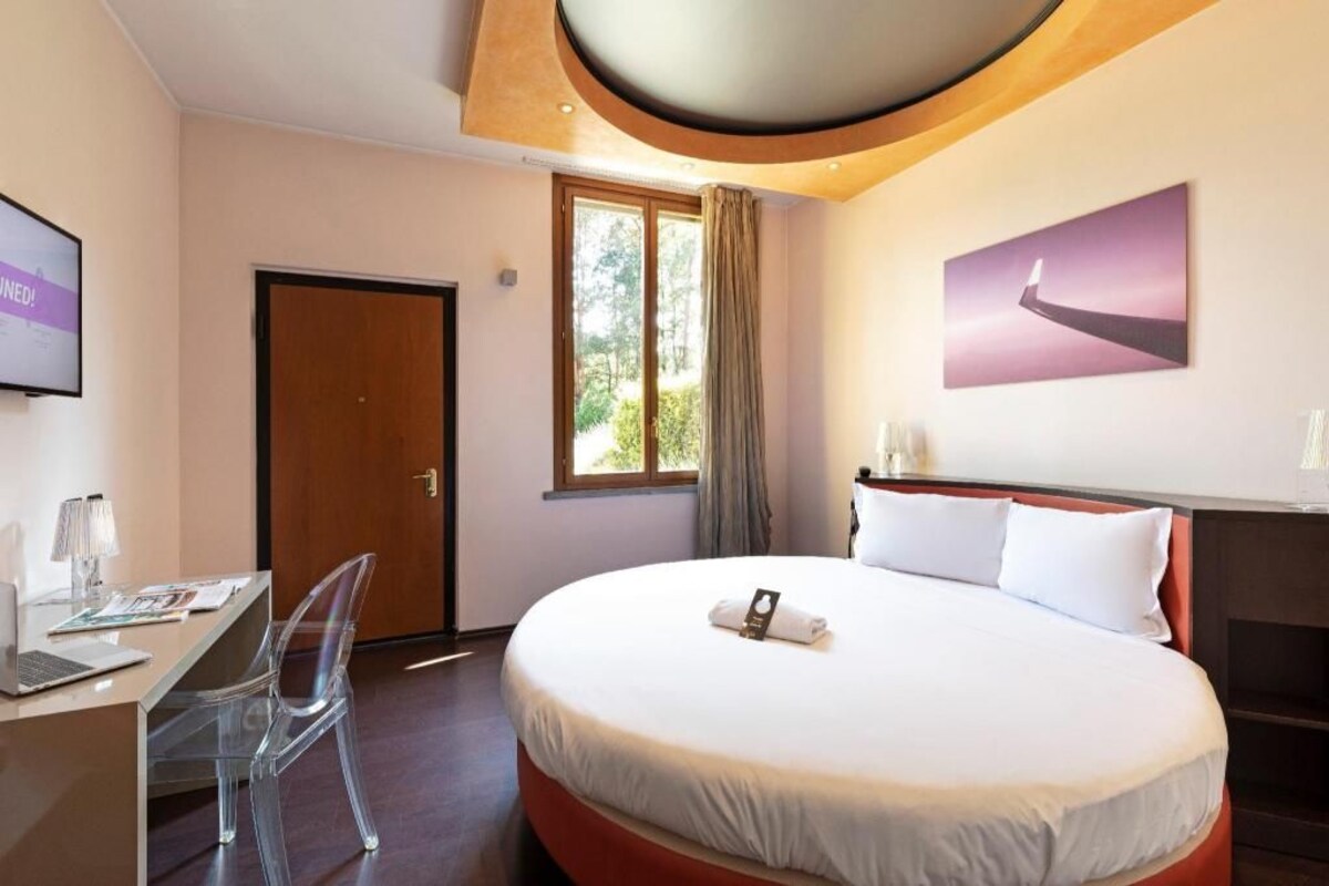 A hotel room with a bed, a desk with a laptop, a chair, a door, a window, and a painting on the wall.
