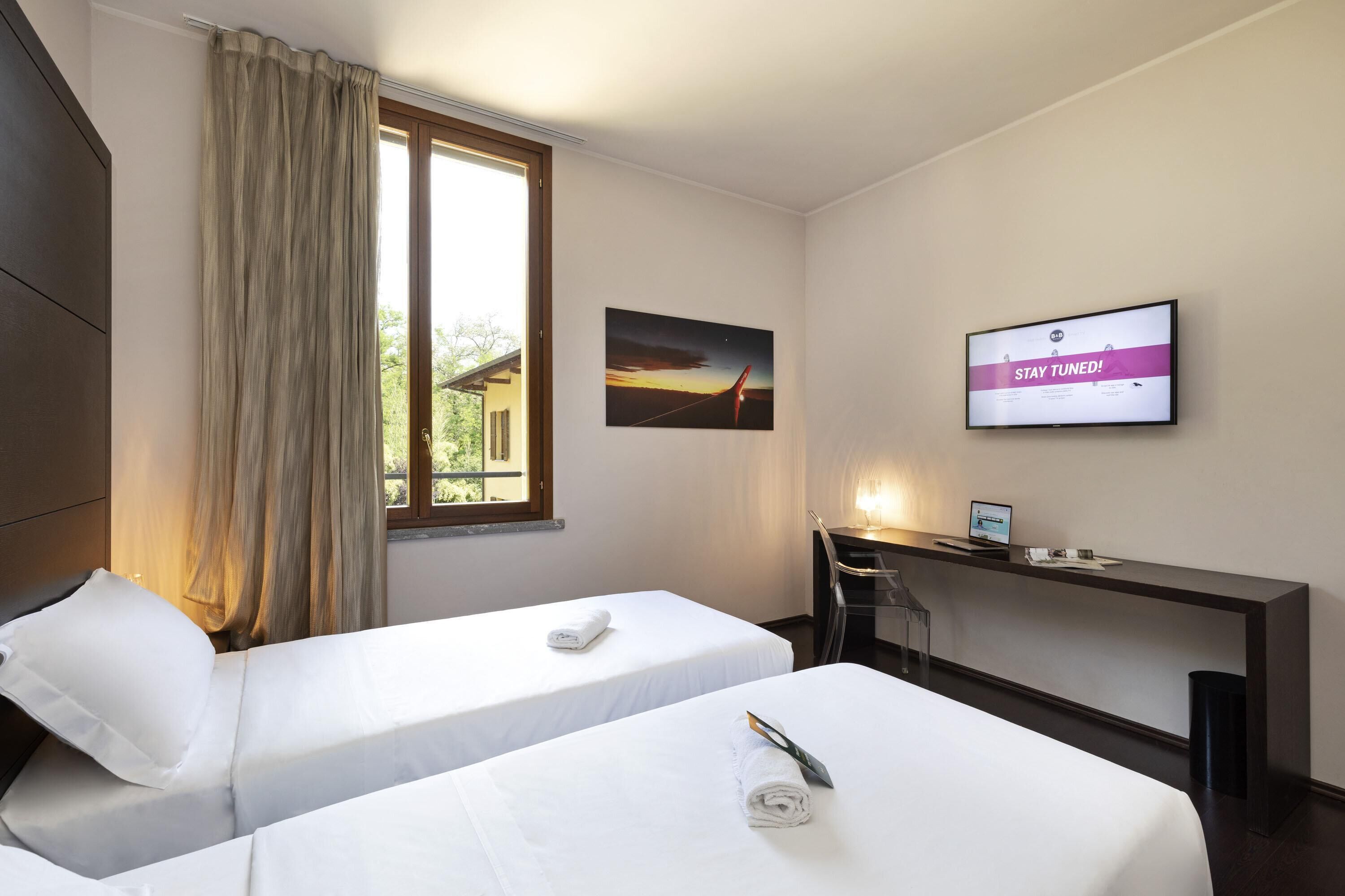 A hotel room with two beds, a desk, a TV, and a window with curtains.