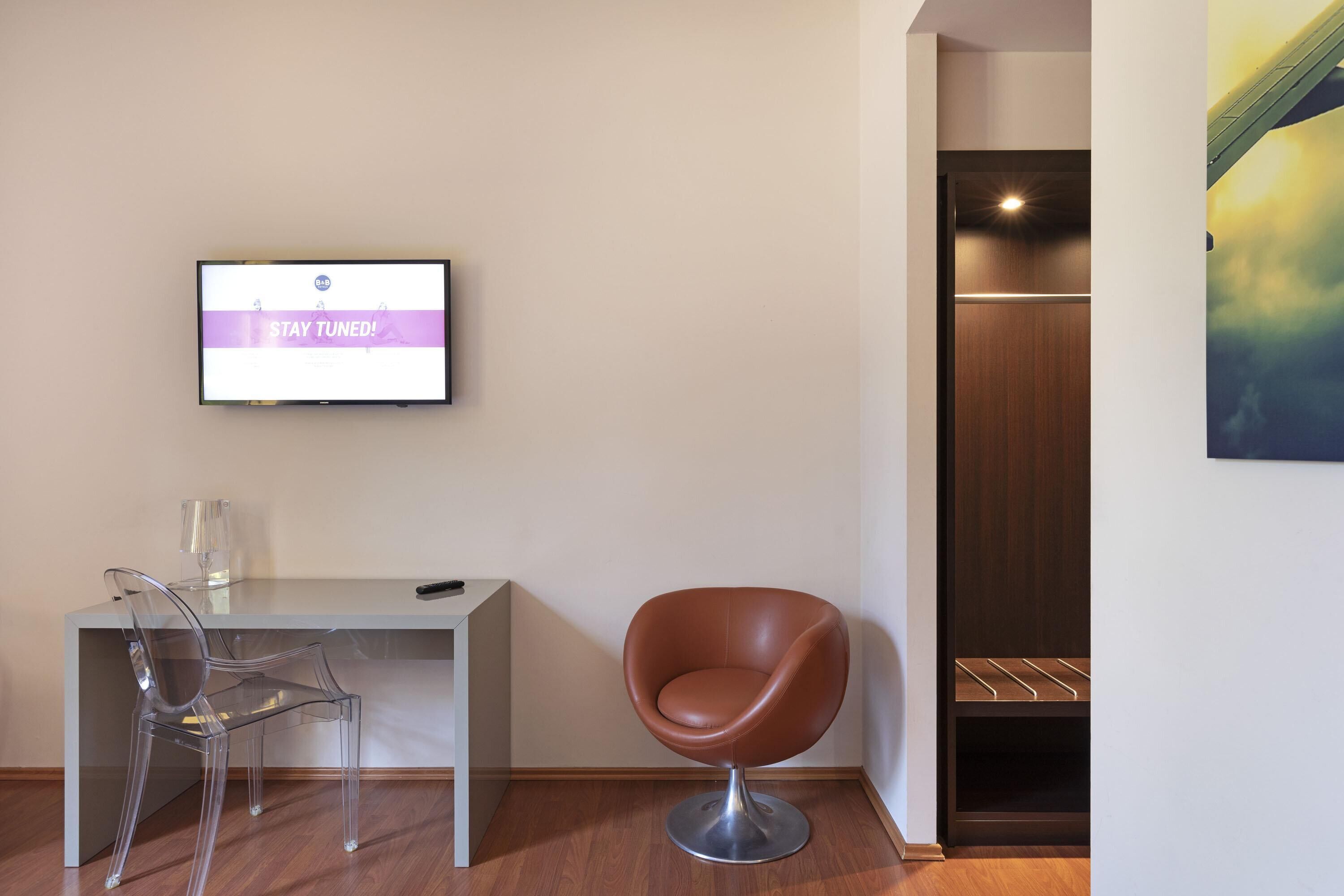 Een moderne kamer met een bureau, twee stoelen, een televisie waarop 'STAY TUNED!' staat en een houten garderobekast.