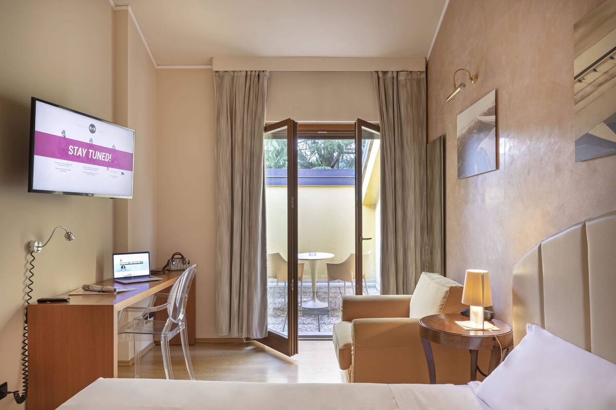 A hotel room with a desk, chair, bed, bedside table, lamp, and a TV displaying a message.