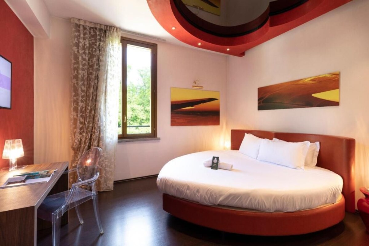 Camera d'albergo con un letto rotondo, una scrivania con una sedia, una finestra con tende e due opere d'arte appese al muro.