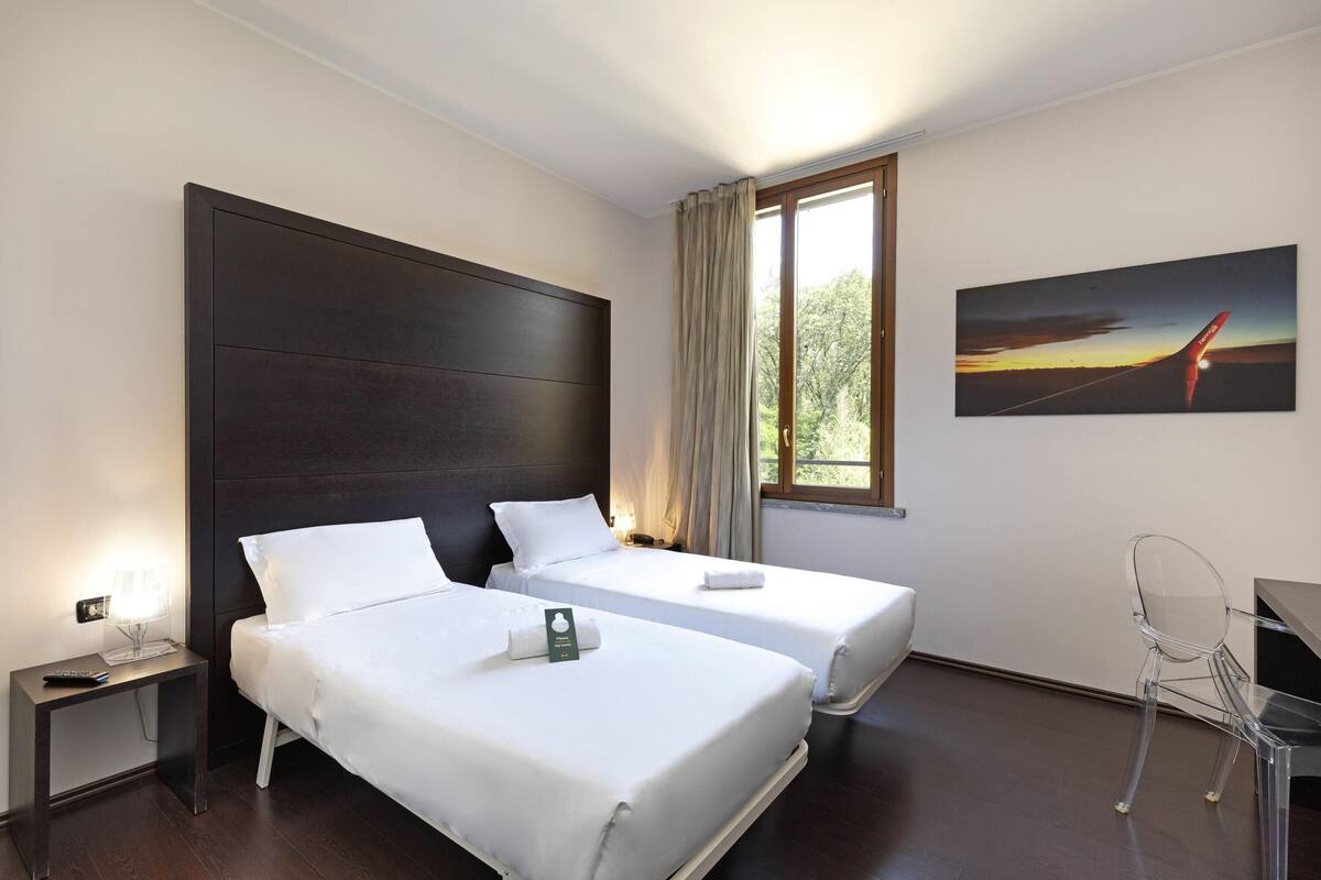 A hotel room with two beds, a desk, a chair, a nightstand, a lamp, and a painting of an airplane.