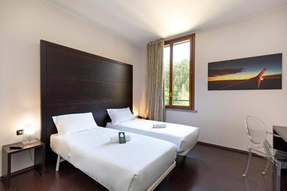 A hotel room with two beds, a desk, a chair, a window with curtains, and a painting on the wall.