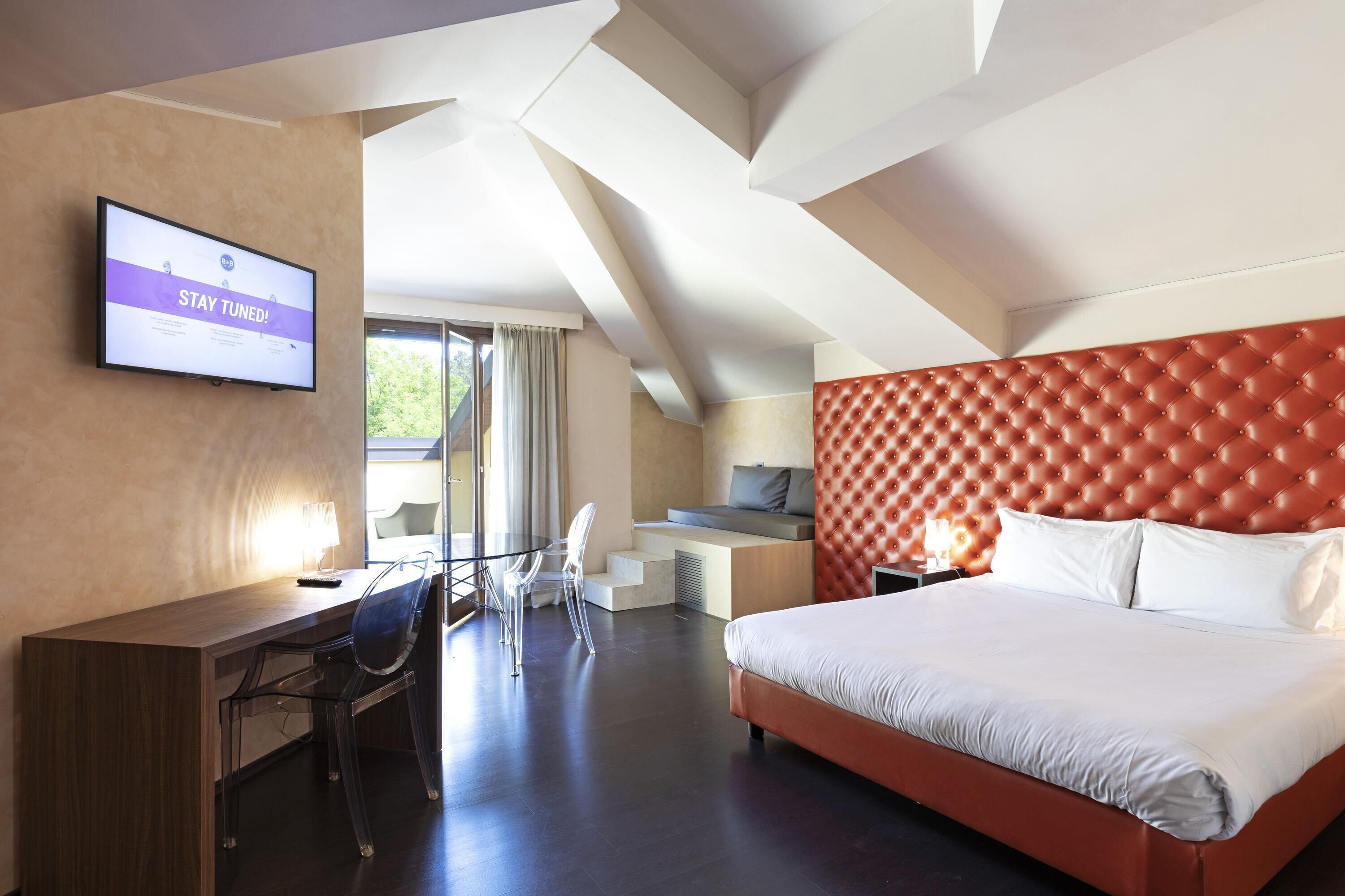 Habitación de hotel con una cama grande, un escritorio y un televisor que muestra la frase 'STAY TUNED'.