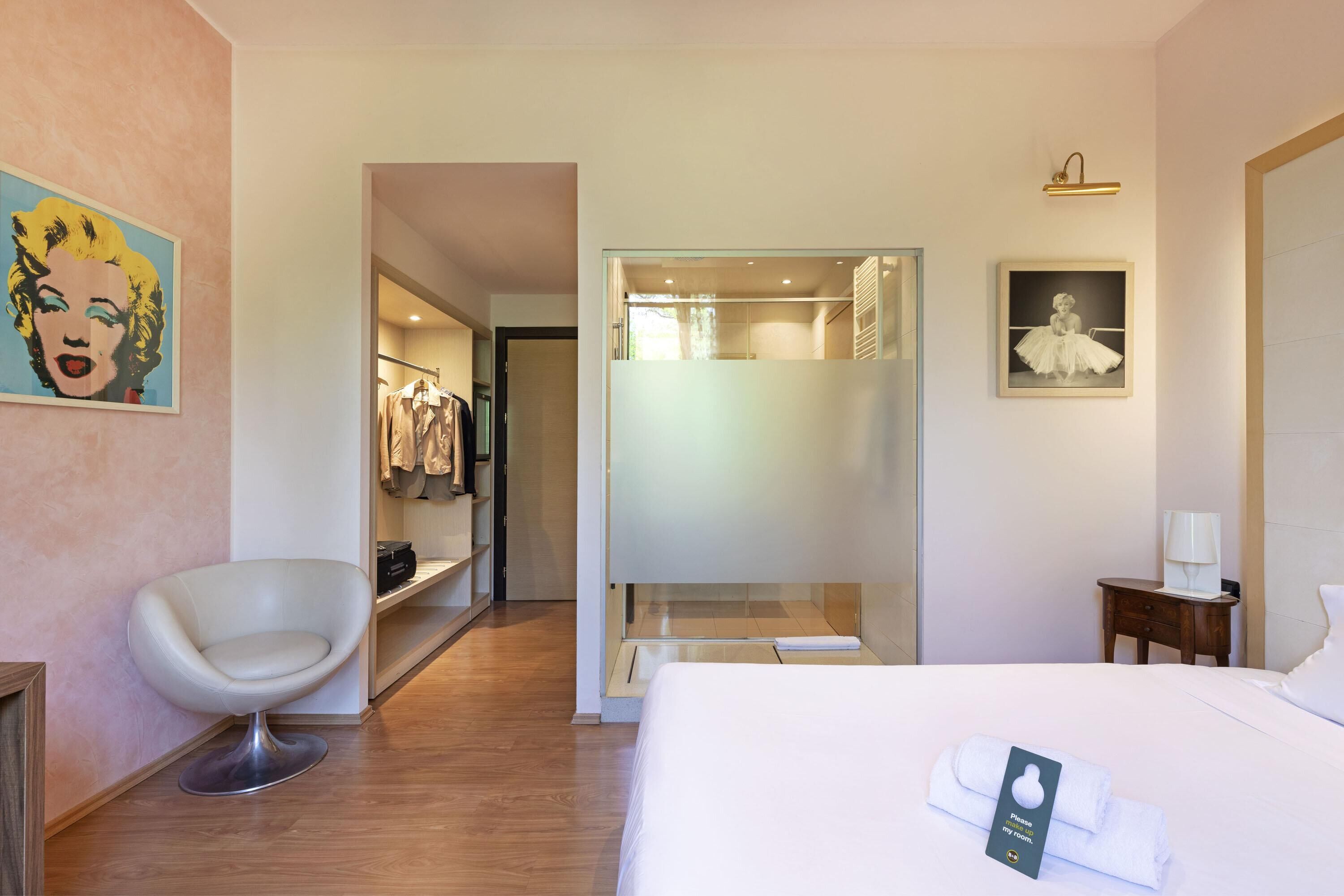Habitación de hotel con cama, una silla, armario y baño visible a través de una puerta de vidrio.