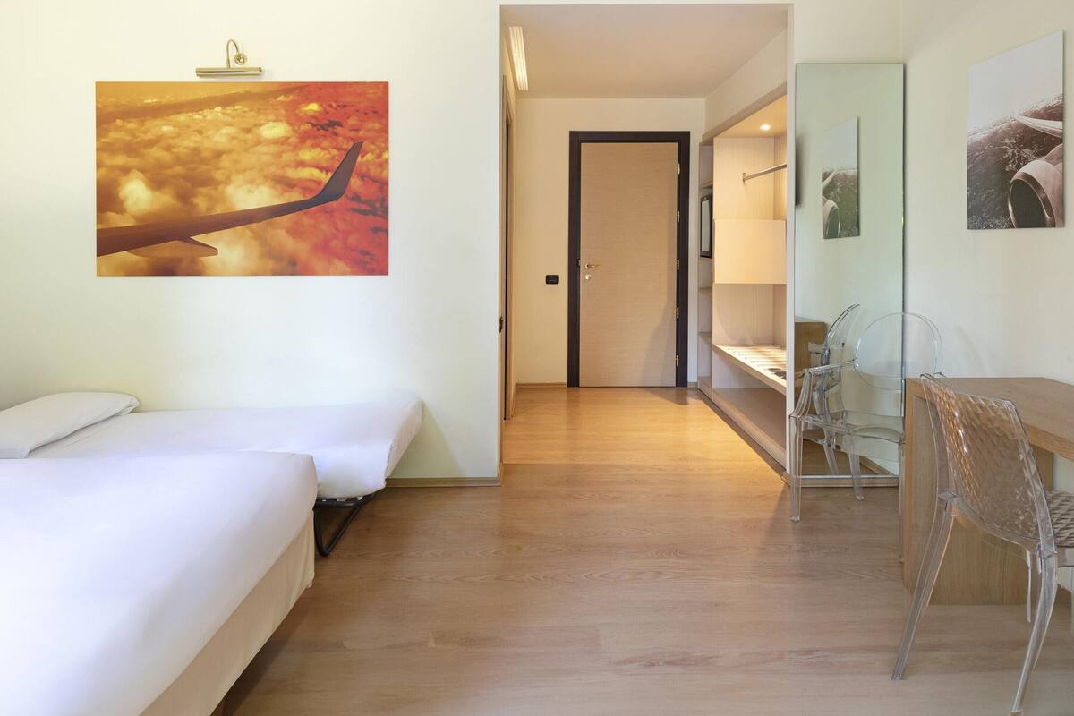 A hotel room with a bed, a desk, and a chair. There is a door leading to another room.
