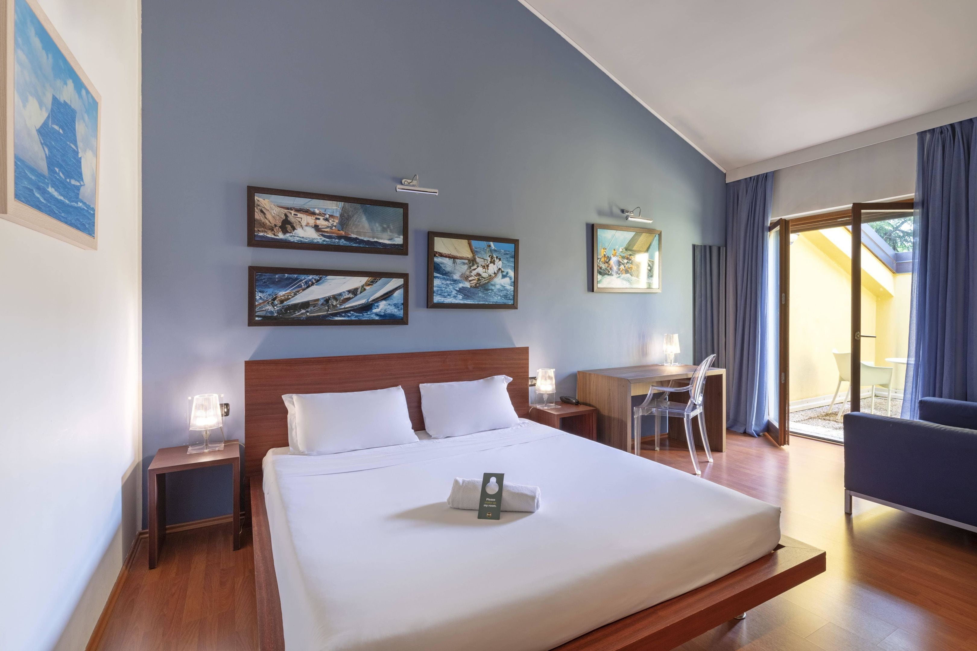 Ein Hotelzimmer mit einem großen Bett, einem Schreibtisch und einem Sessel. An der Wand befinden sich Gemälde und ein Fenster mit Vorhängen.