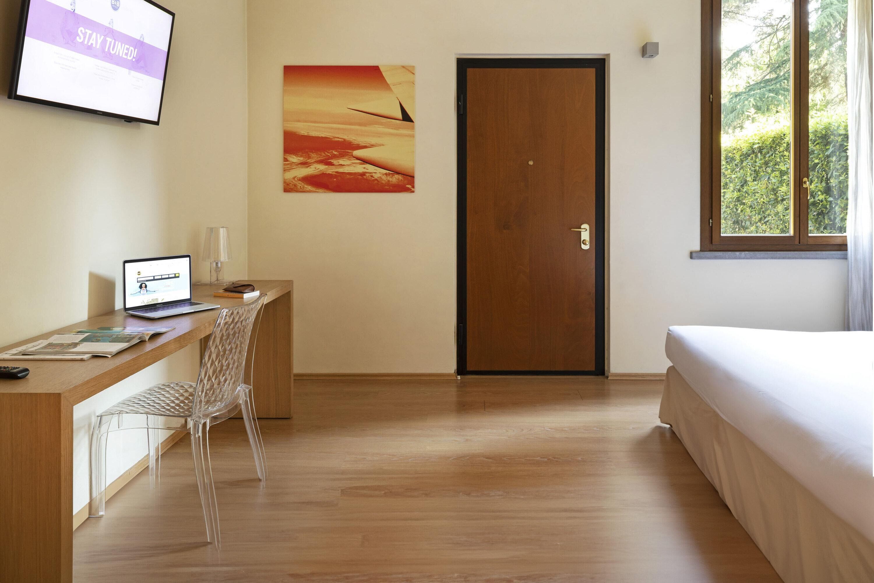 A hotel room with a wooden desk, a laptop, a clear chair, a bed, a door, a window with a view of greenery, and a wall-mounted TV displaying "STAY TUNED!".