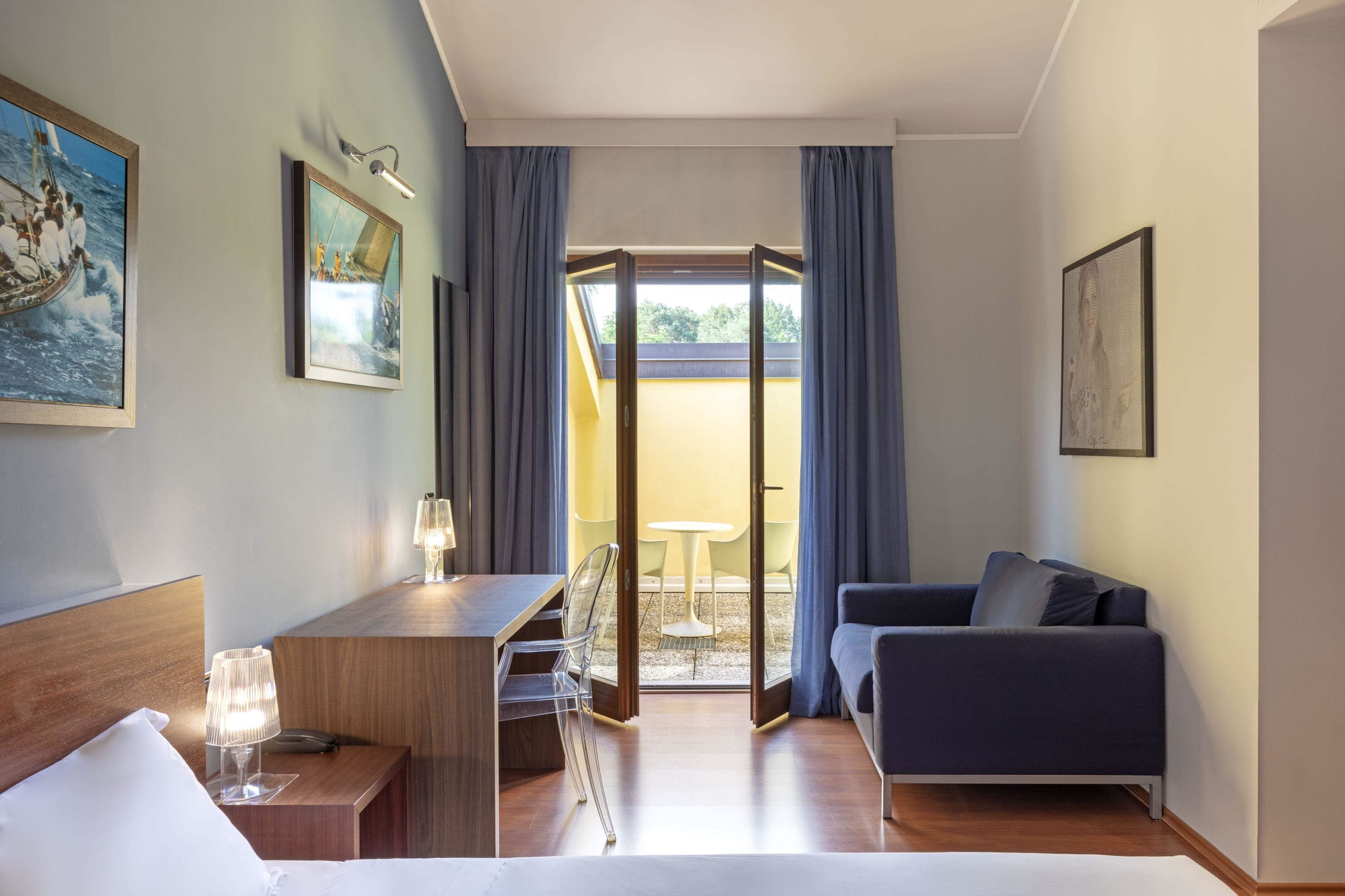 Ein Hotelzimmer mit Bett, Schreibtisch mit Lampe, Stuhl, Sofa und Balkon mit Tisch und Stühlen.
